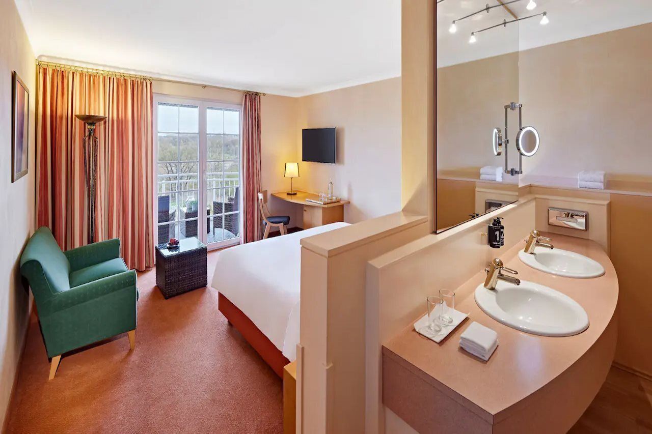 Ett hotellrum med en säng, en grön fåtölj, ett skrivbord med en lampa, en tv, två handfat och en stor spegel.