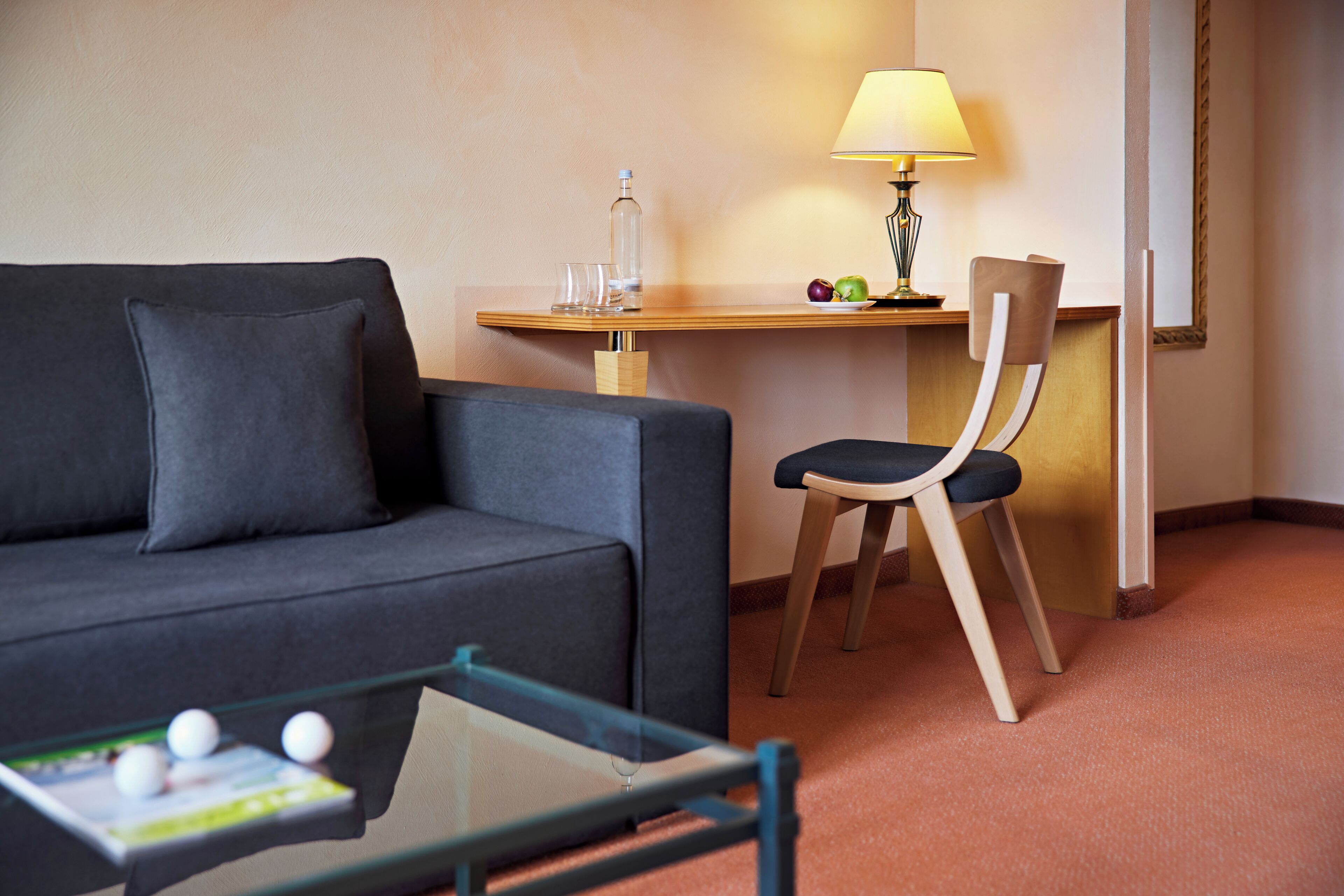 A modern hotel room with a grey sofa, a wooden side table with a lamp, a glass coffee table, and a chair.