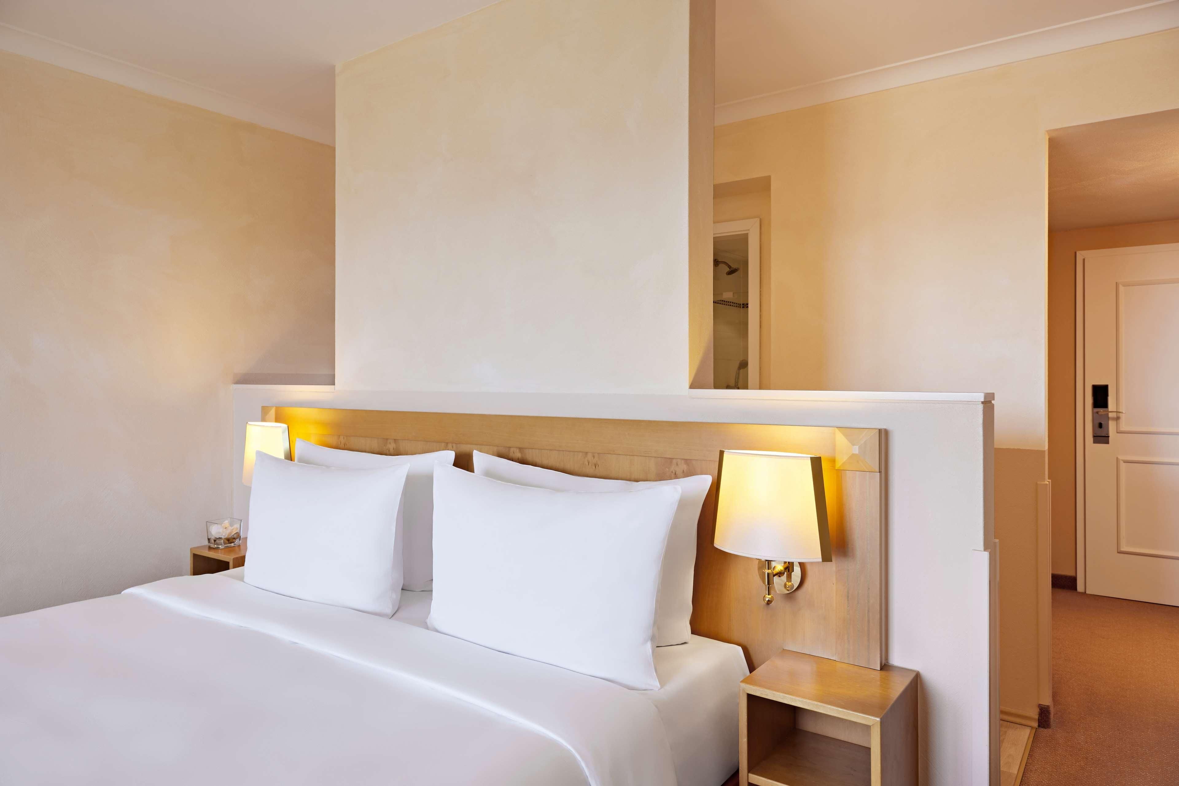 A hotel room with a large bed, two bedside tables, a nightstand, a wall-mounted lamp, and a mirror above the bed.