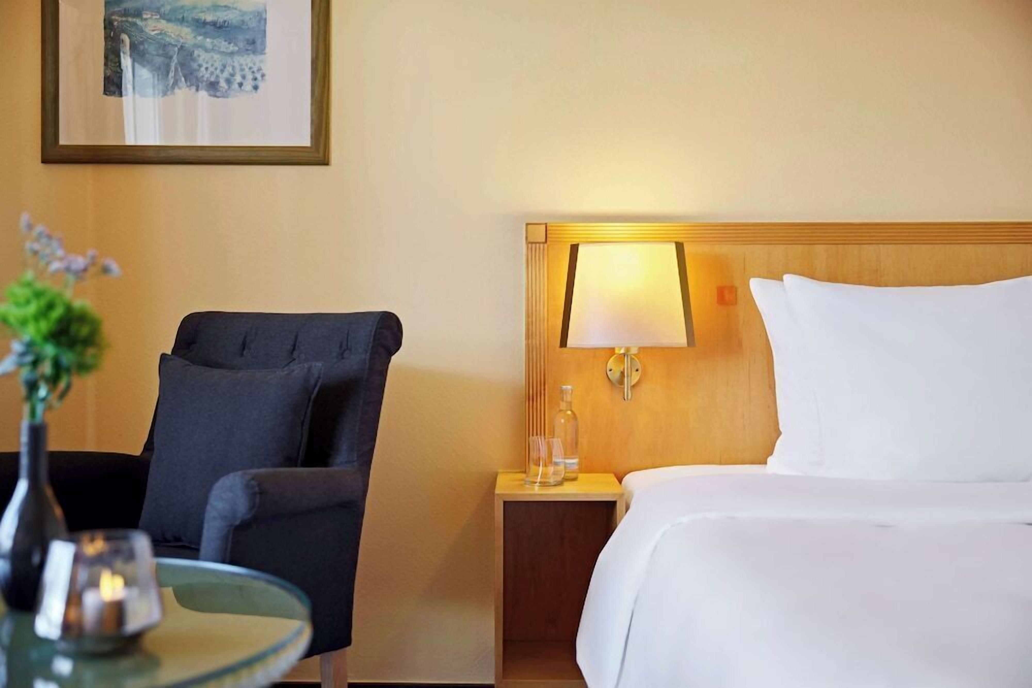 Een hotelkamer met een bed, een nachtkastje met een lamp, een stoel en een kleine tafel met een vaas en kaars.