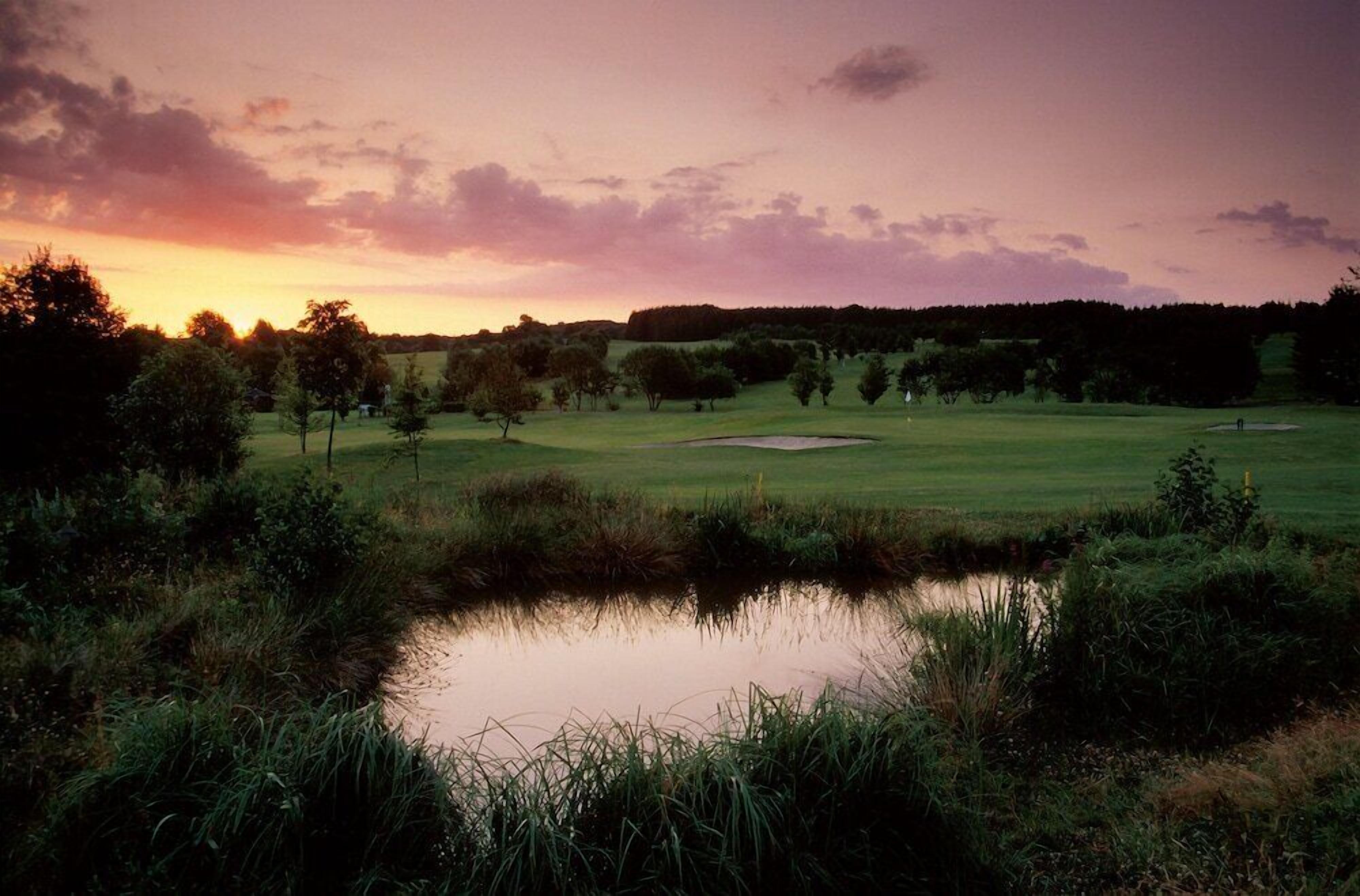 Ein Golfplatz bei Sonnenuntergang mit einem Teich im Vordergrund und Bäumen in der Umgebung.