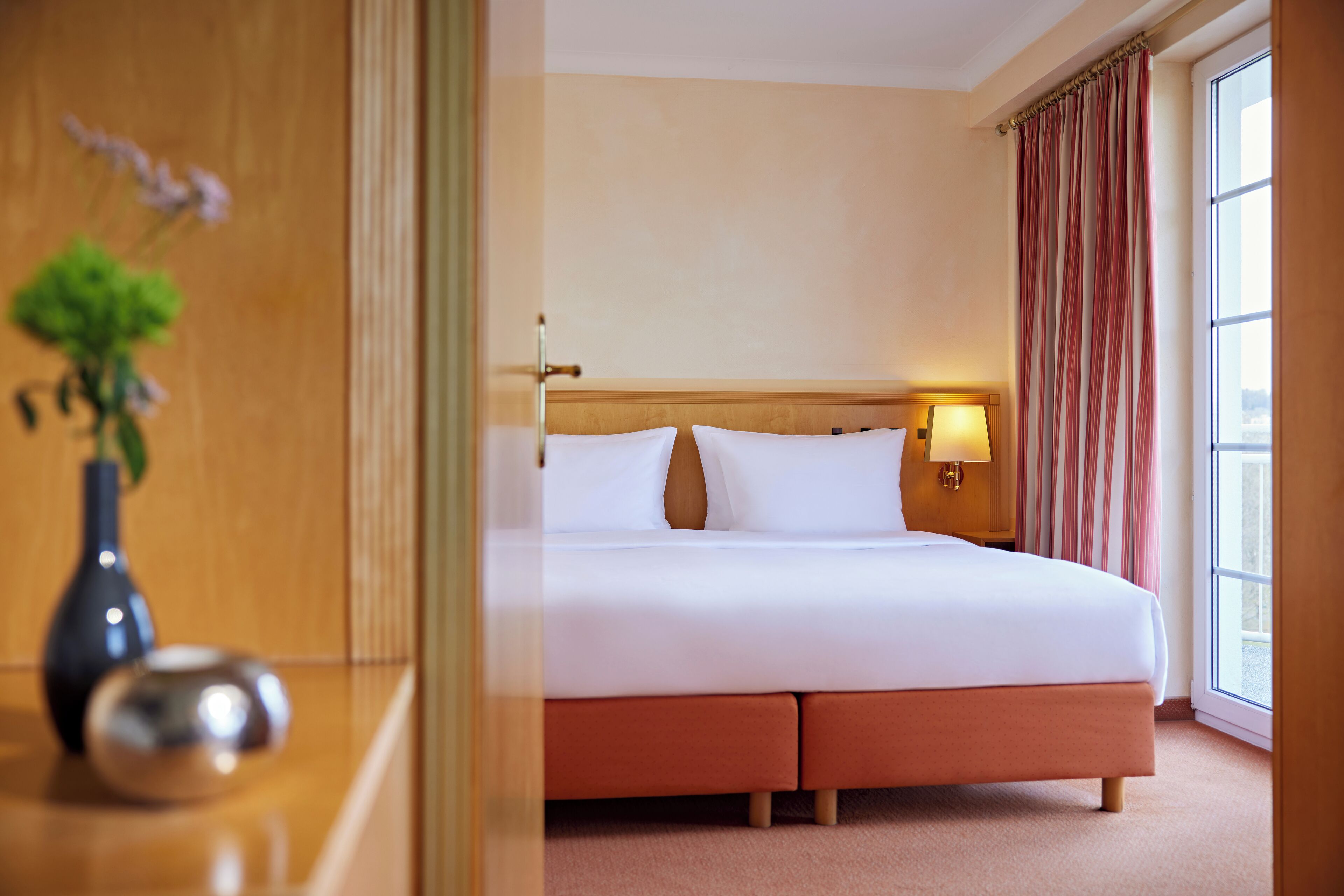 A hotel room with a bed, a nightstand, a lamp, a vase with flowers, and a window with curtains.