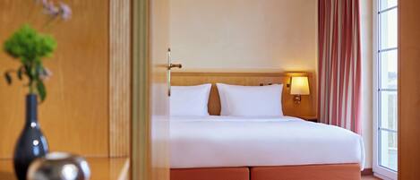 A hotel room with a bed, a nightstand, a lamp, a vase with flowers, and a window with curtains.