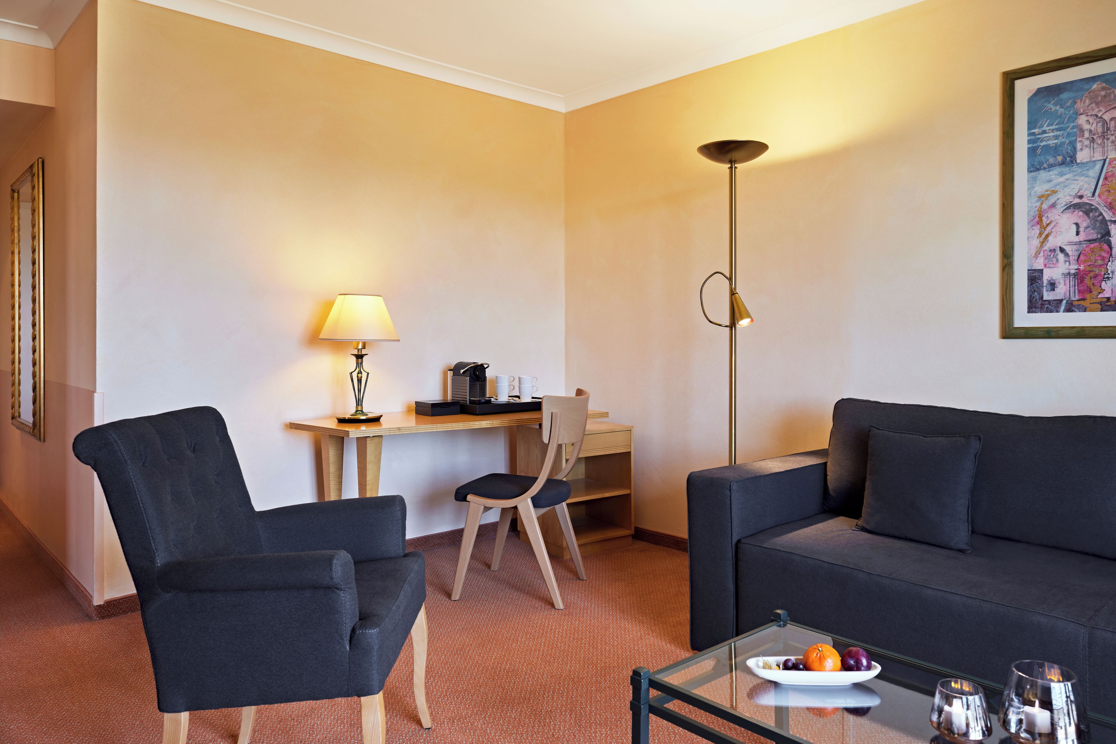 A modern living room with a sofa, two armchairs, a coffee table with fruit, a lamp, and a desk with a chair.