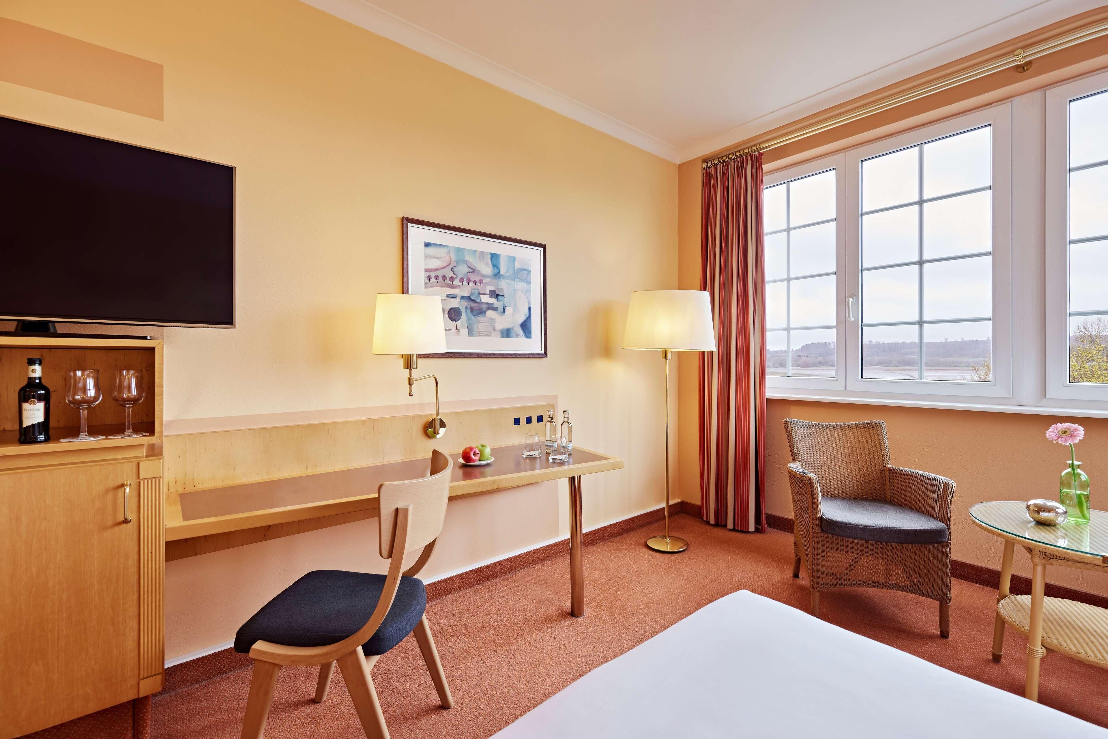 Habitación de hotel con cama, escritorio con silla, televisión, ventana con cortinas y una pequeña mesa con un florero.