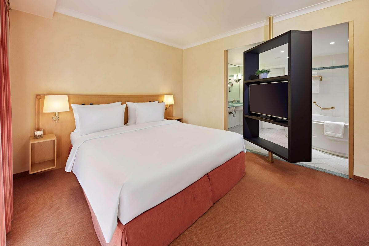 A hotel room with a large bed, bedside lamps, a TV, and a bathroom visible through an open door.