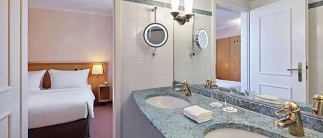 A hotel bathroom with two sinks, a large mirror, and a bed in the adjacent room.