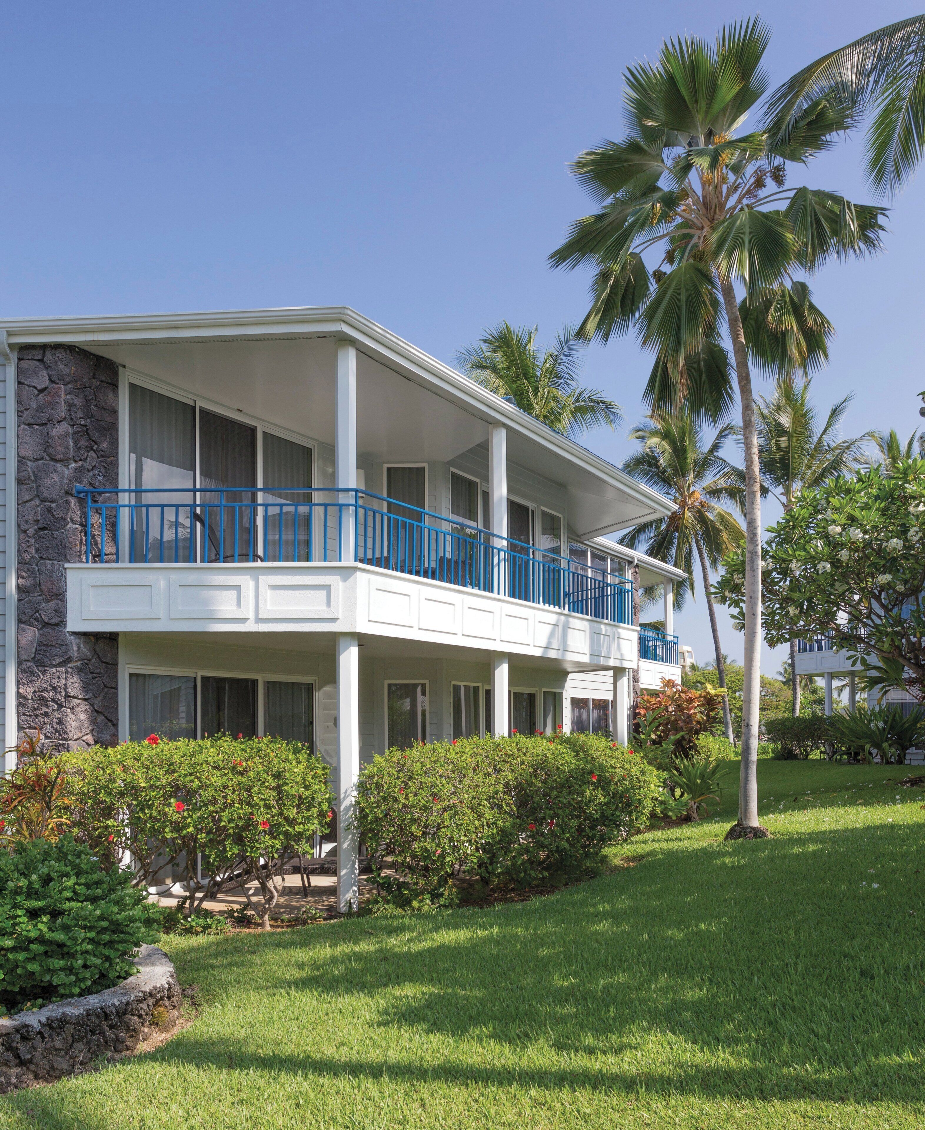A two-story building with a balcony, stone facade, and lush greenery.