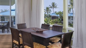 A dining area with a large table and chairs, large windows with curtains, and a view of palm trees and buildings outside. - Holua Resort (Kailua-Kona)