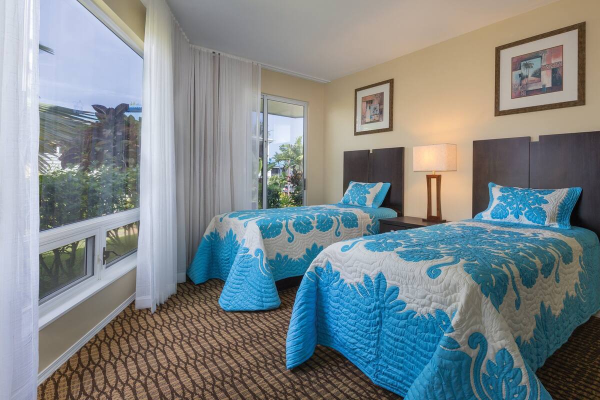 Une chambre d’hôtel avec deux lits, une grande fenêtre avec des rideaux, une vue sur la verdure et un tableau encadré au mur.