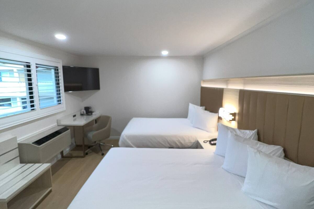A hotel room with two beds, a desk, a chair, a TV, and a window with blinds.