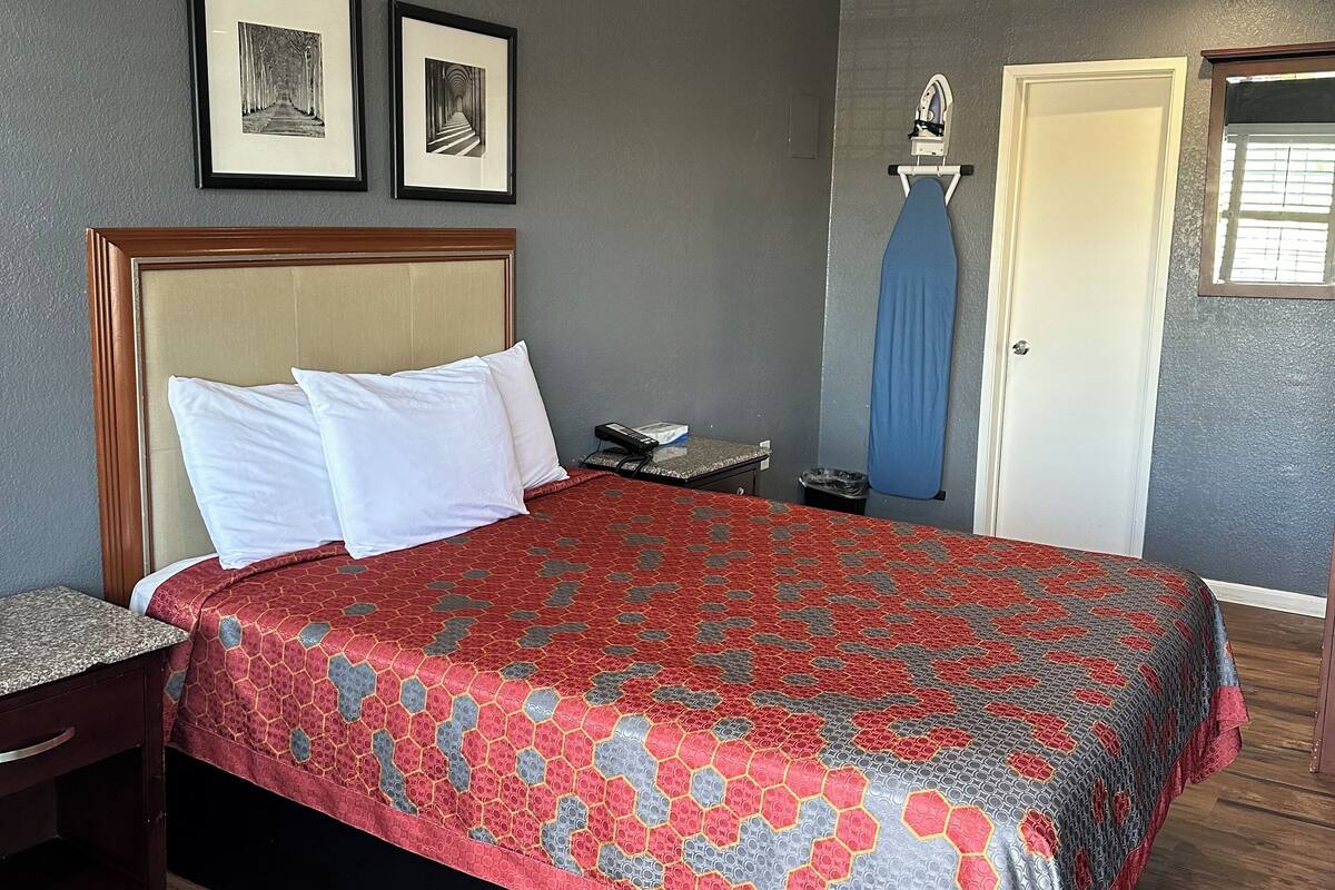 A neatly made bed with a patterned bedspread, a wooden nightstand, a framed picture, a wall-mounted ironing board, and a door with a window.