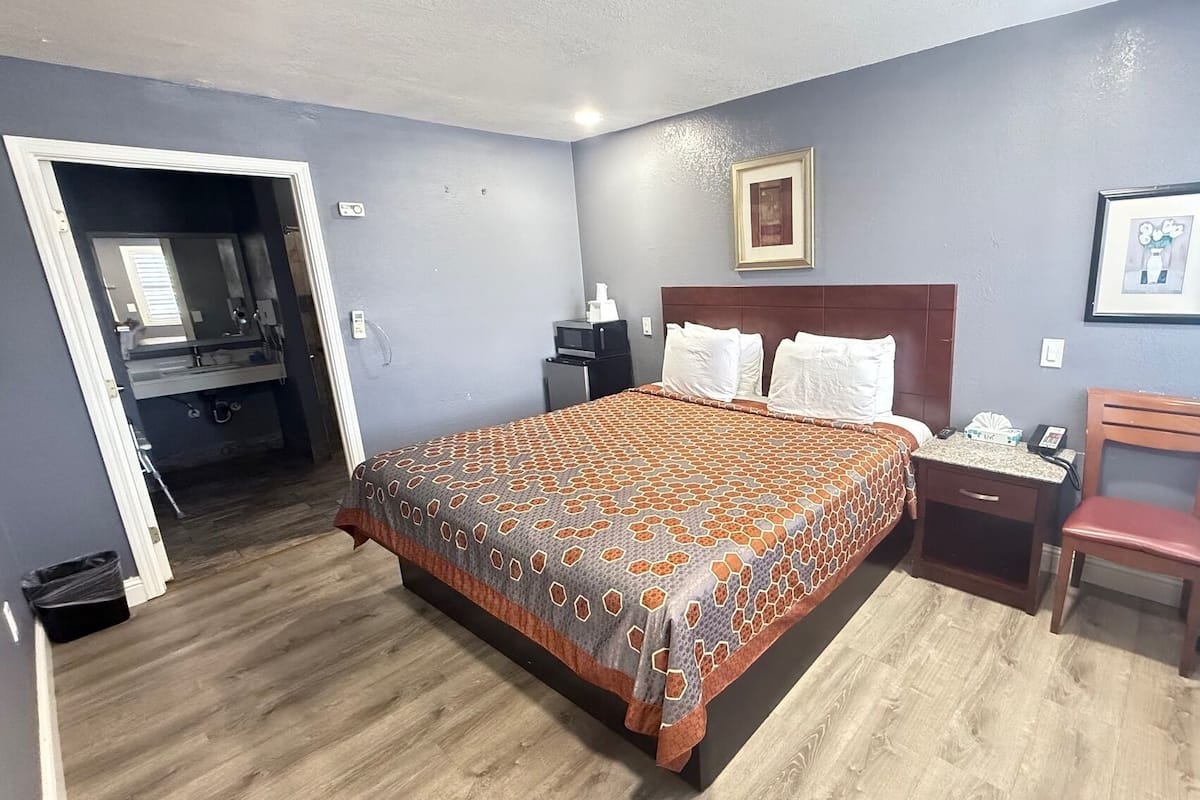 A hotel room with a large bed, a wooden nightstand, a chair, a desk, and a bathroom visible through an open door.