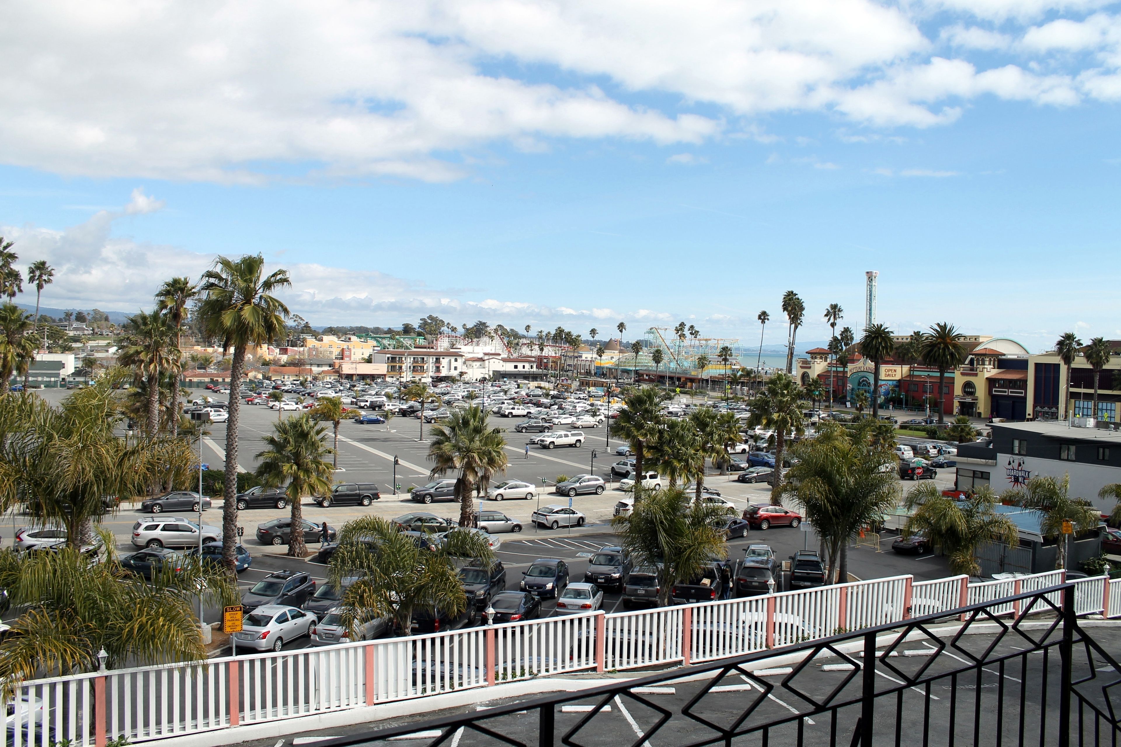 A parking lot with multiple cars and palm trees, surrounded by buildings and a clear sky.