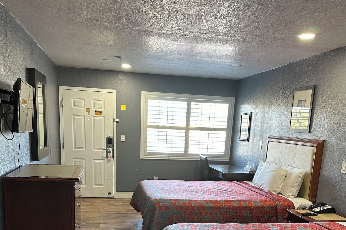 A hotel room with two beds, a desk, a chair, a dresser, and a window with blinds.