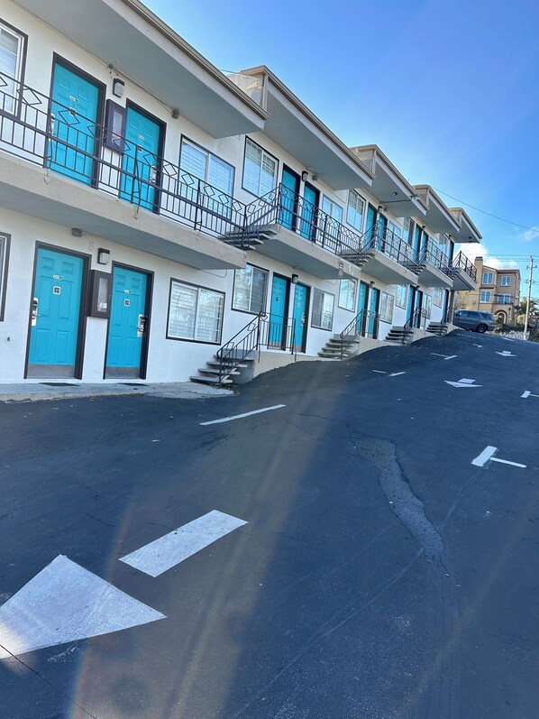 Un hotel de varios pisos con puertas azules y balcones, cielo despejado y un estacionamiento con flechas direccionales.