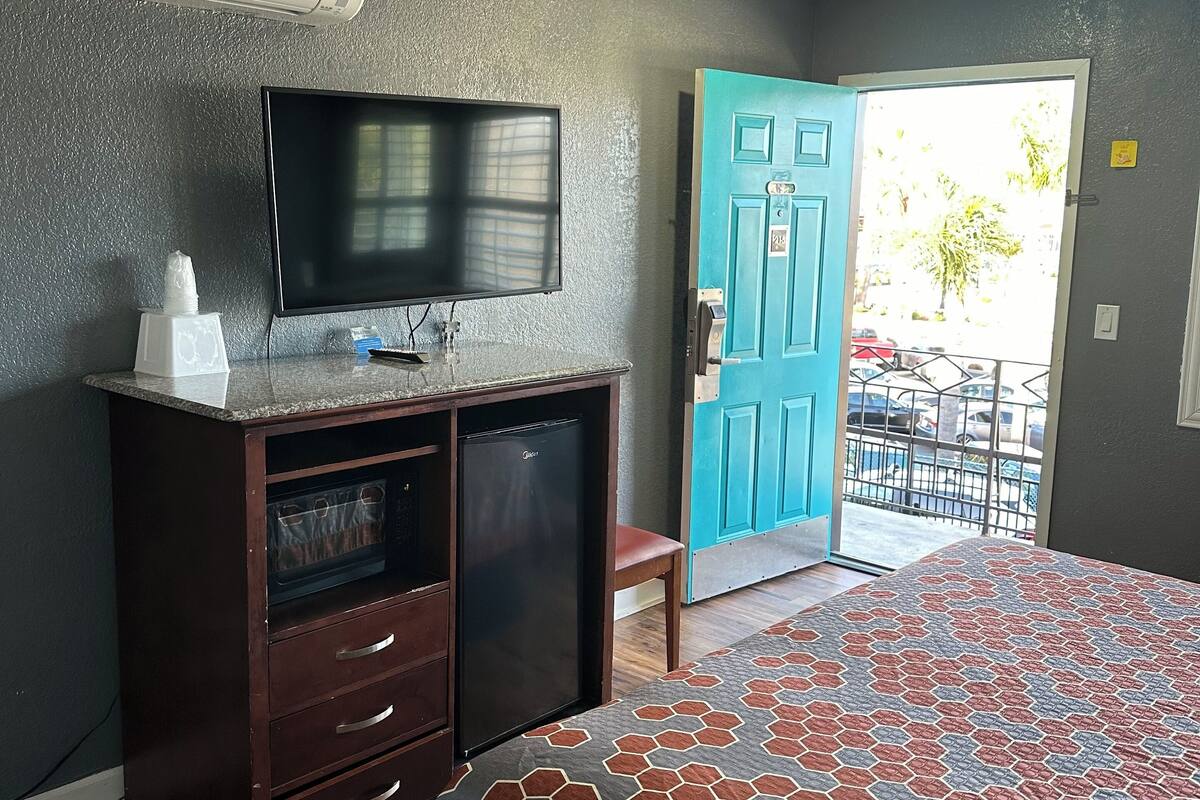 A hotel room with a bed, a TV, a mini-fridge, a wooden dresser, and a door leading outside.