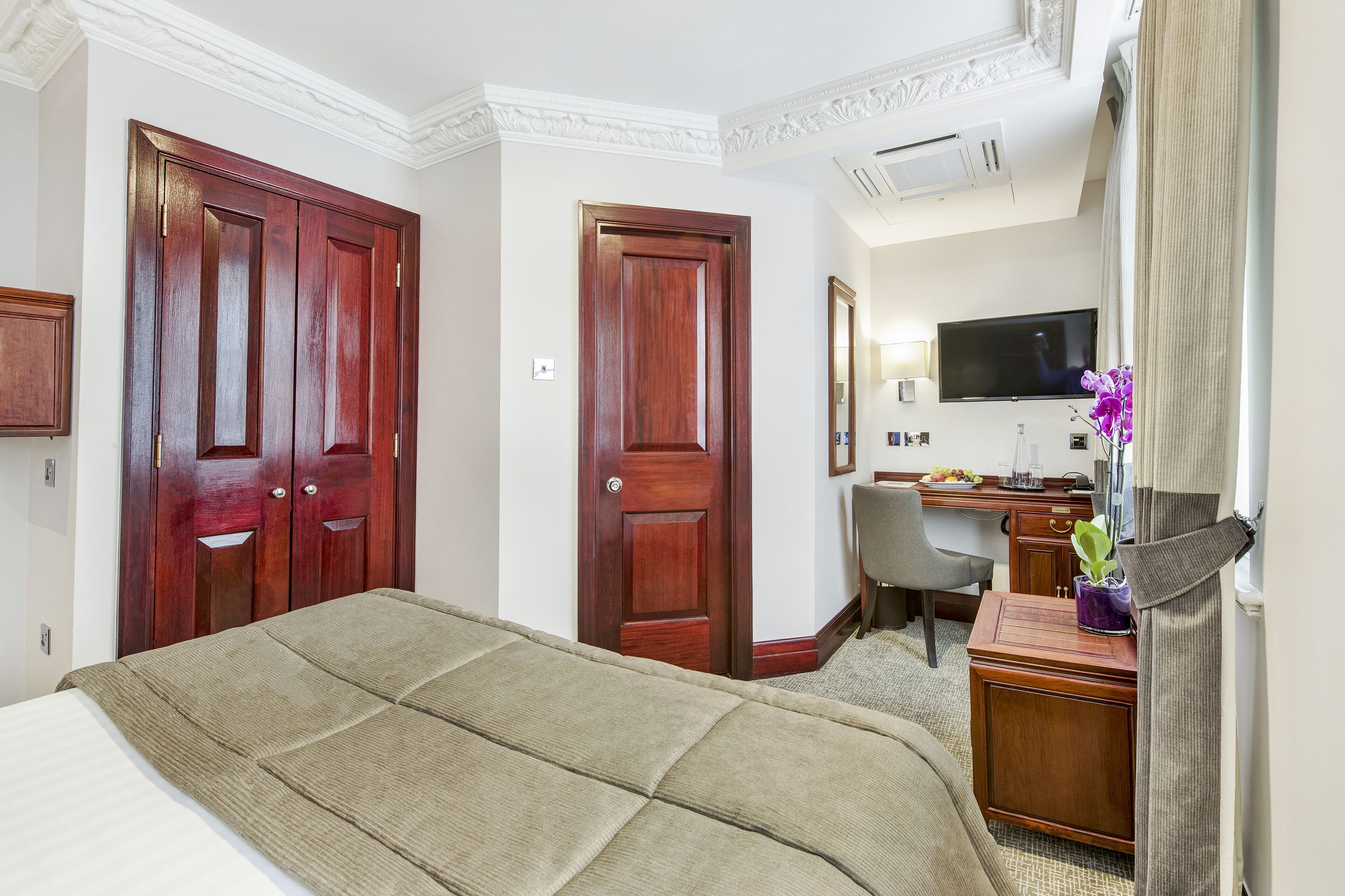 A hotel room with a large bed, a desk with a chair, a television, and two wooden doors.