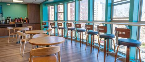 A restaurant interior with wooden tables and chairs, large windows with a view of buildings, and a counter with condiments.