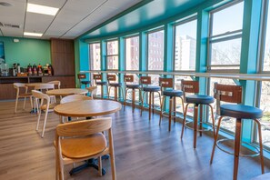 L’intérieur d’un restaurant est aménagé avec des tables et des chaises en bois, de grandes fenêtres offrant une vue sur les bâtiments, et un comptoir où sont disposés les condiments.