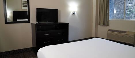 Una camera d'albergo con un letto, una TV su una cassettiera, una lampada a parete, una finestra con tende e un condizionatore d'aria.