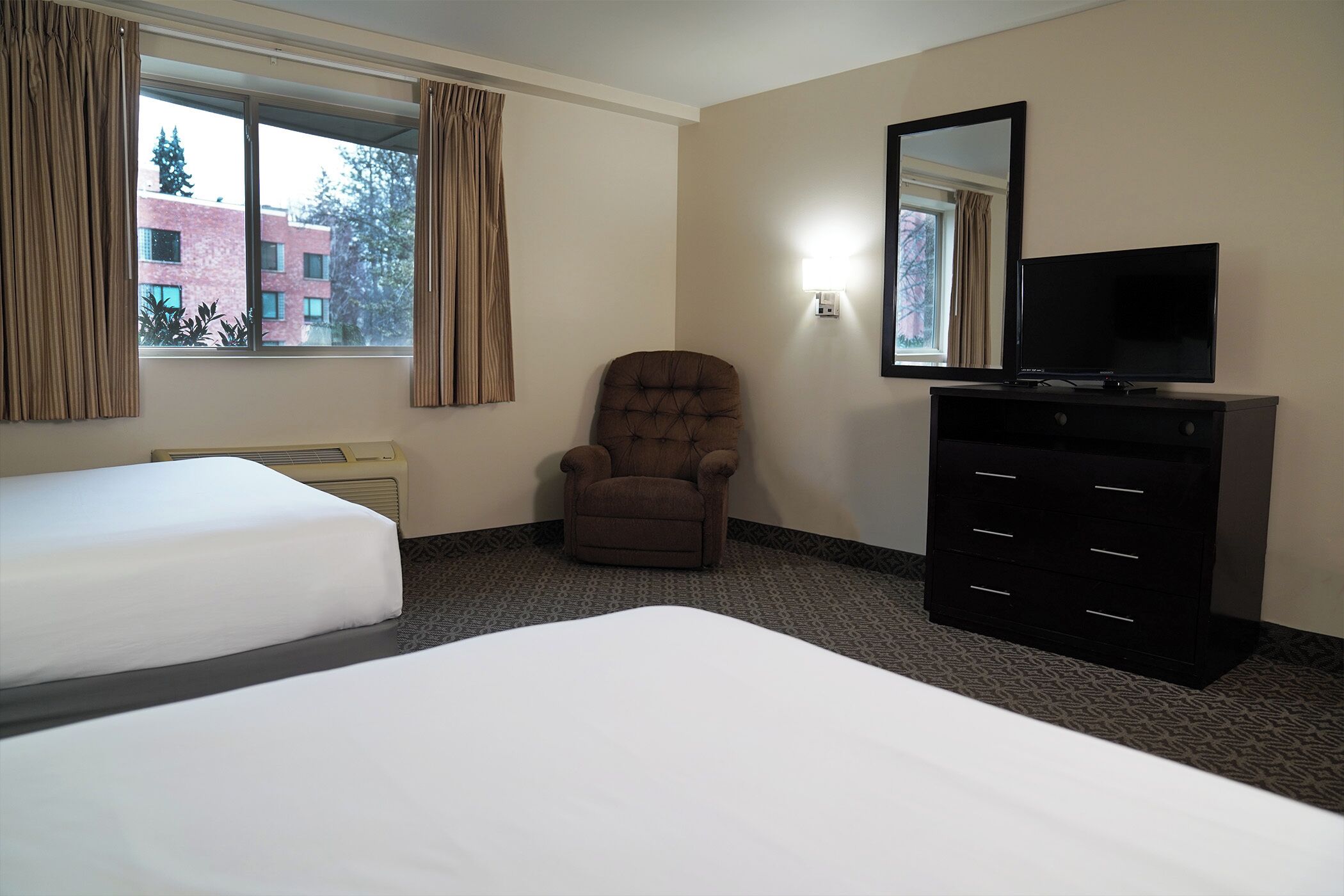 A hotel room with two beds, a brown armchair, a black dresser, and a large window with curtains.