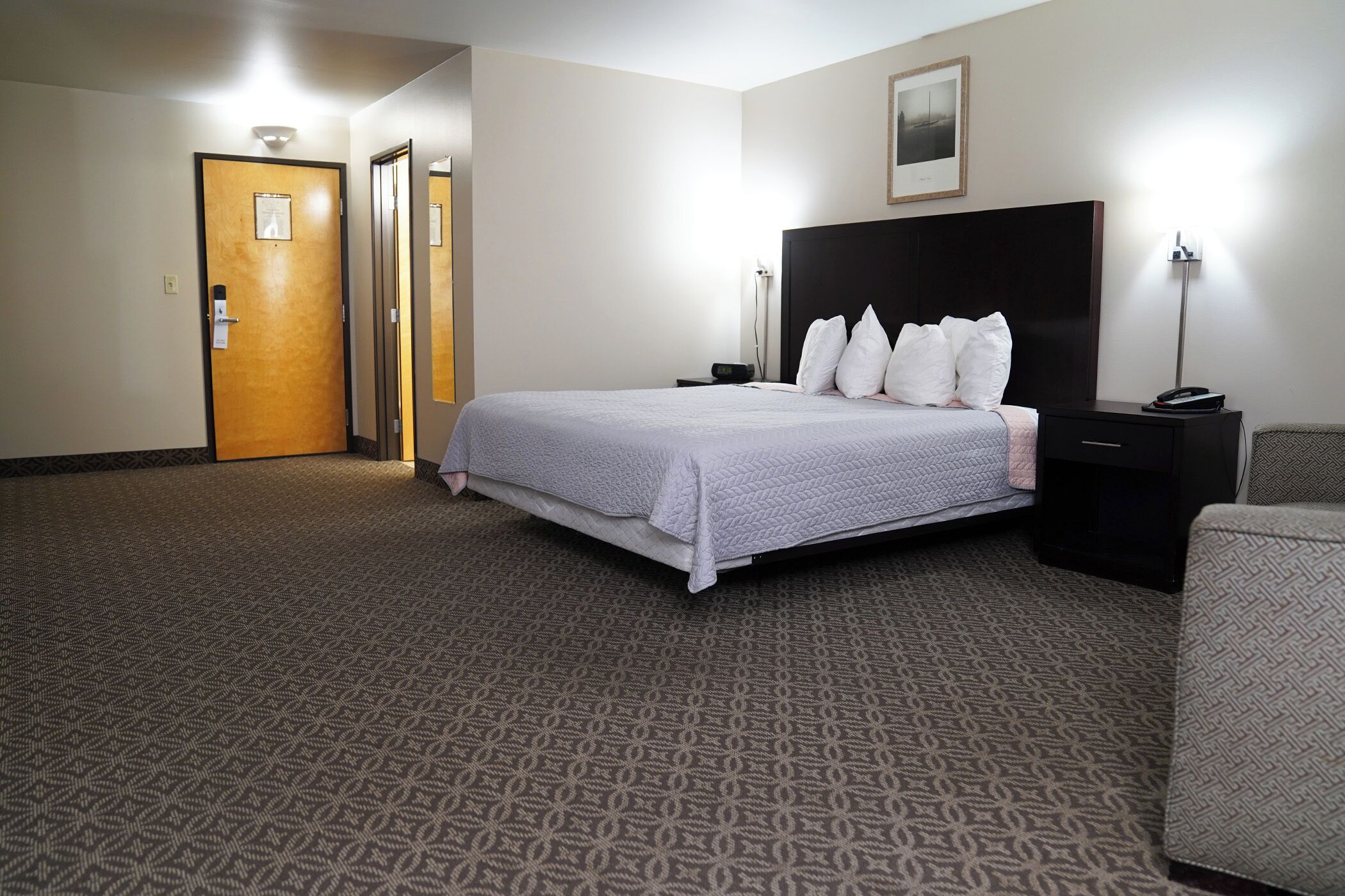 Habitación de hotel con una cama grande, una mesita de noche, una silla y una puerta con luz encima.
