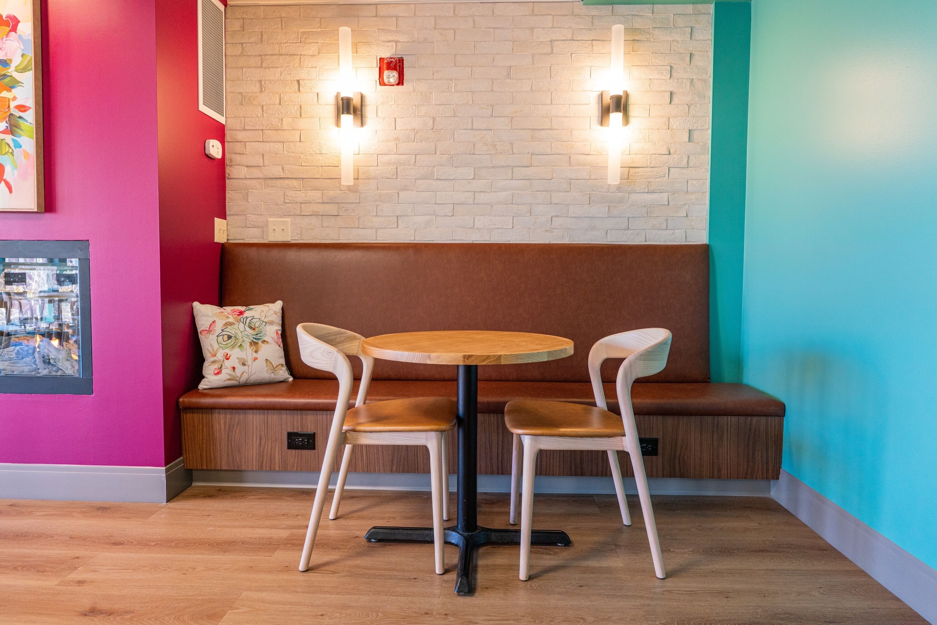 A seating area with a round table and two chairs, a bench with a floral pillow, and a brick wall with two wall-mounted lights.