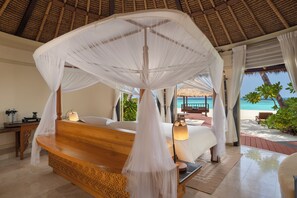 A bedroom with a canopy bed, a wooden bench, a bedside table, and a view of the beach. - Banyan Tree Vabbinfaru (Vabbinfaru)
