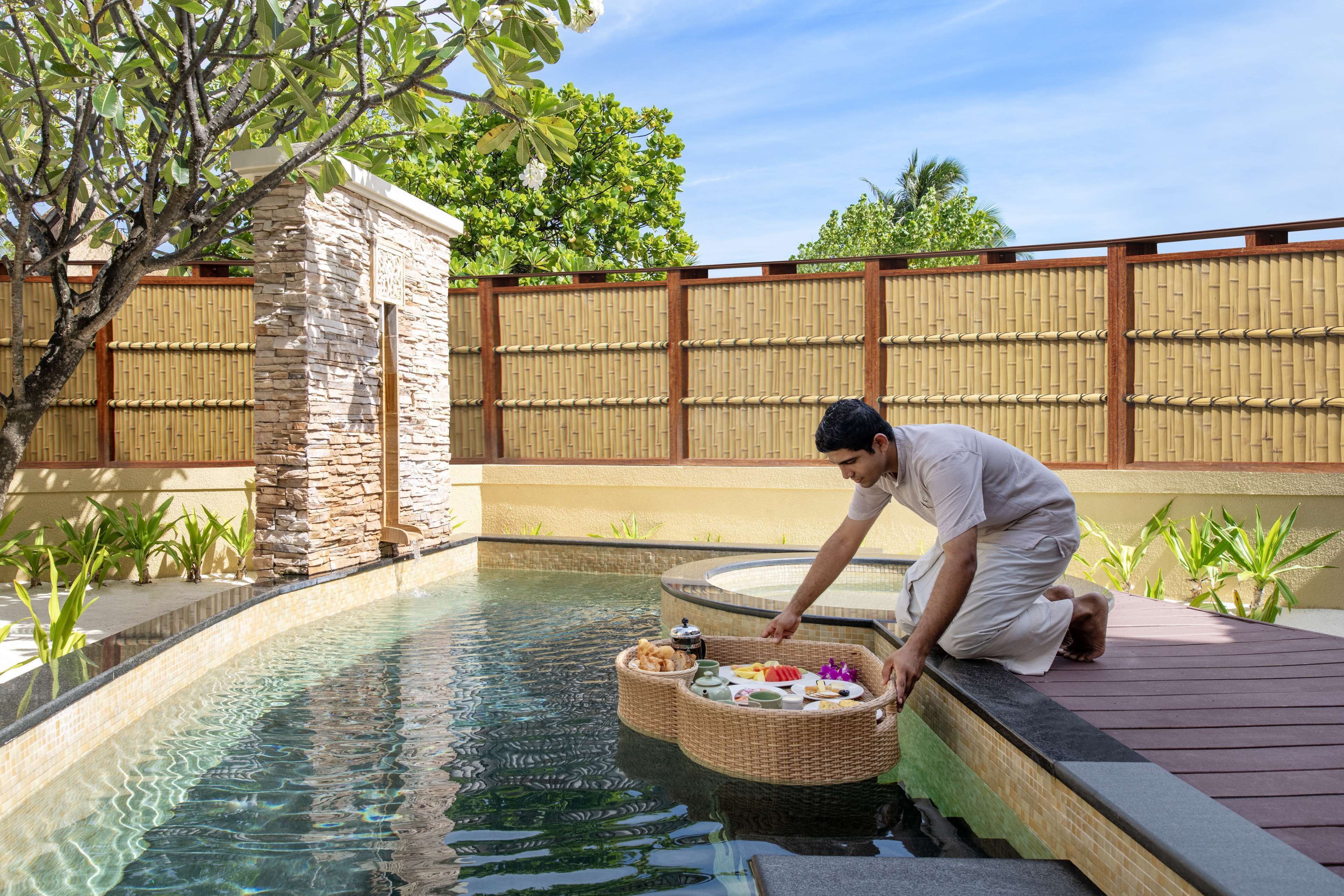 Zona junto a la piscina con pared de piedra, valla de bambú y una persona sirviendo comida desde una cesta.