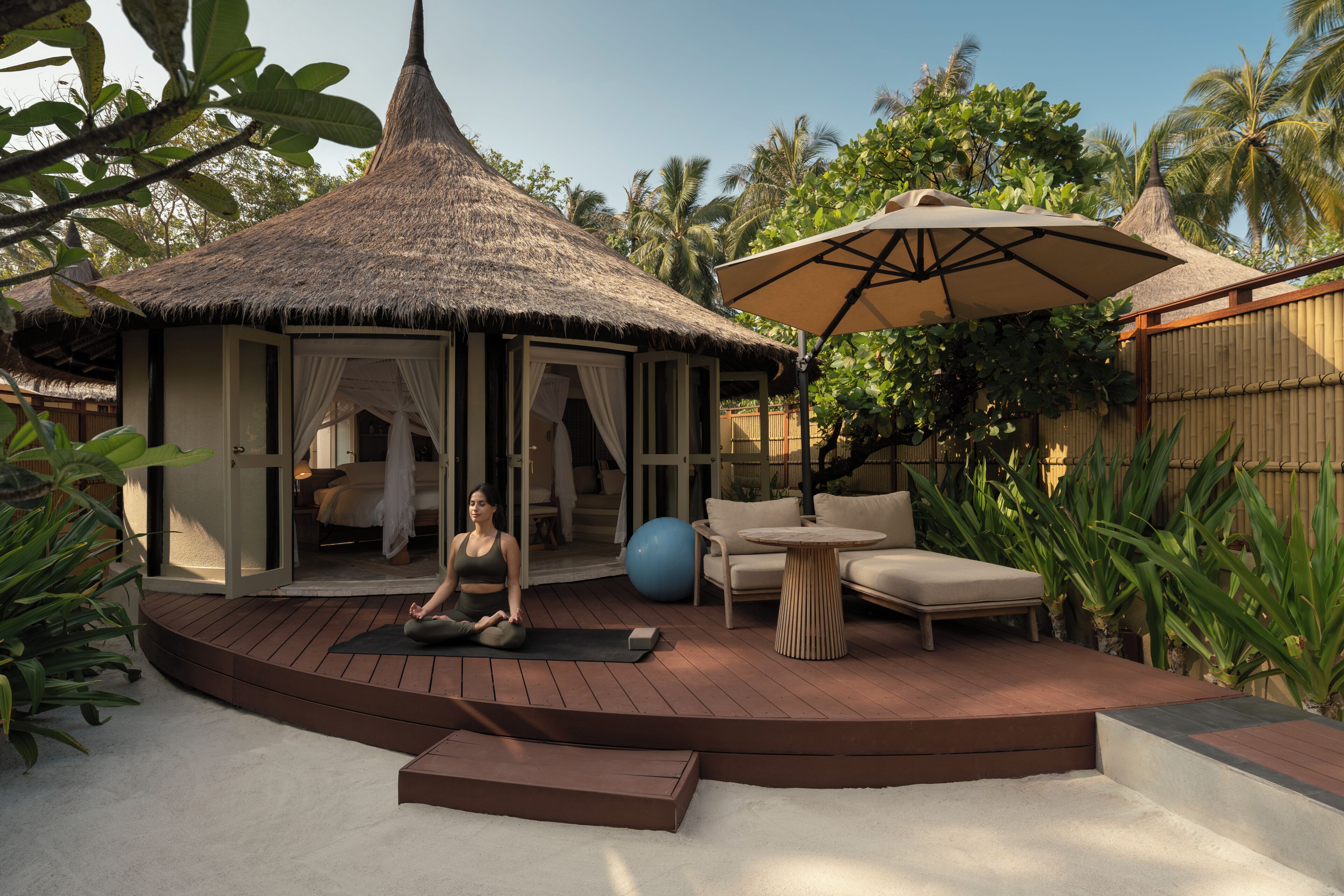 A thatched-roof structure with a covered entrance, a yoga mat, a blue exercise ball, and a round table with a chair.