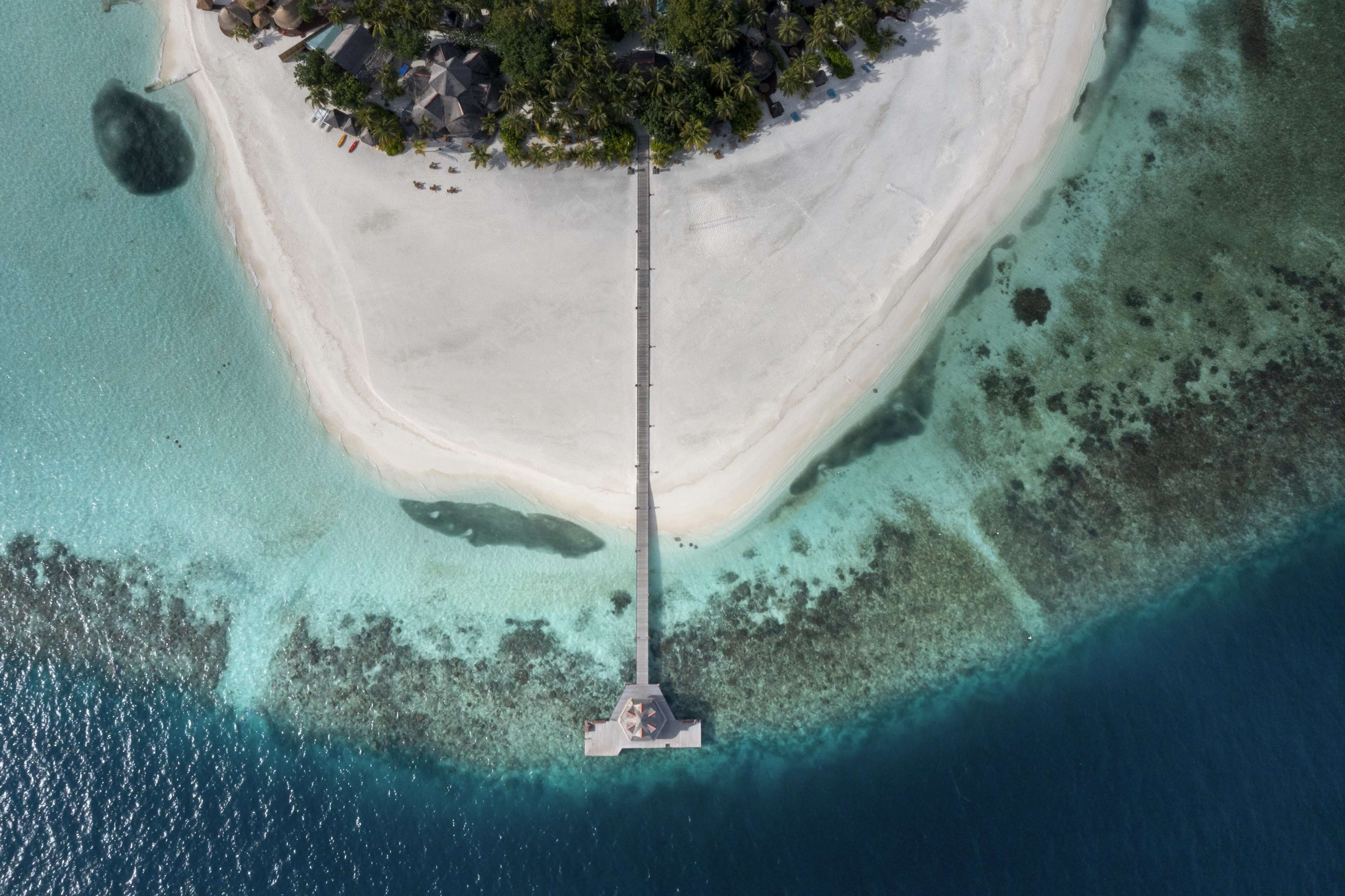 An aerial view of a beach with a long pier extending into the water, surrounded by clear turquoise waters and a small island with trees and buildings.