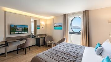 A hotel room with a bed, a desk, a chair, a TV, and a window with a view of the sea.