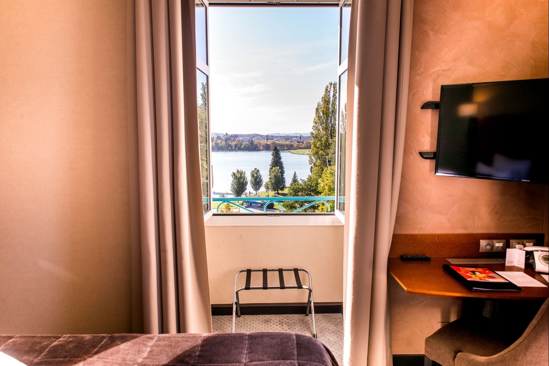 A hotel room with a large window offering a view of a lake and park, a bed, a desk with a TV, and a chair.