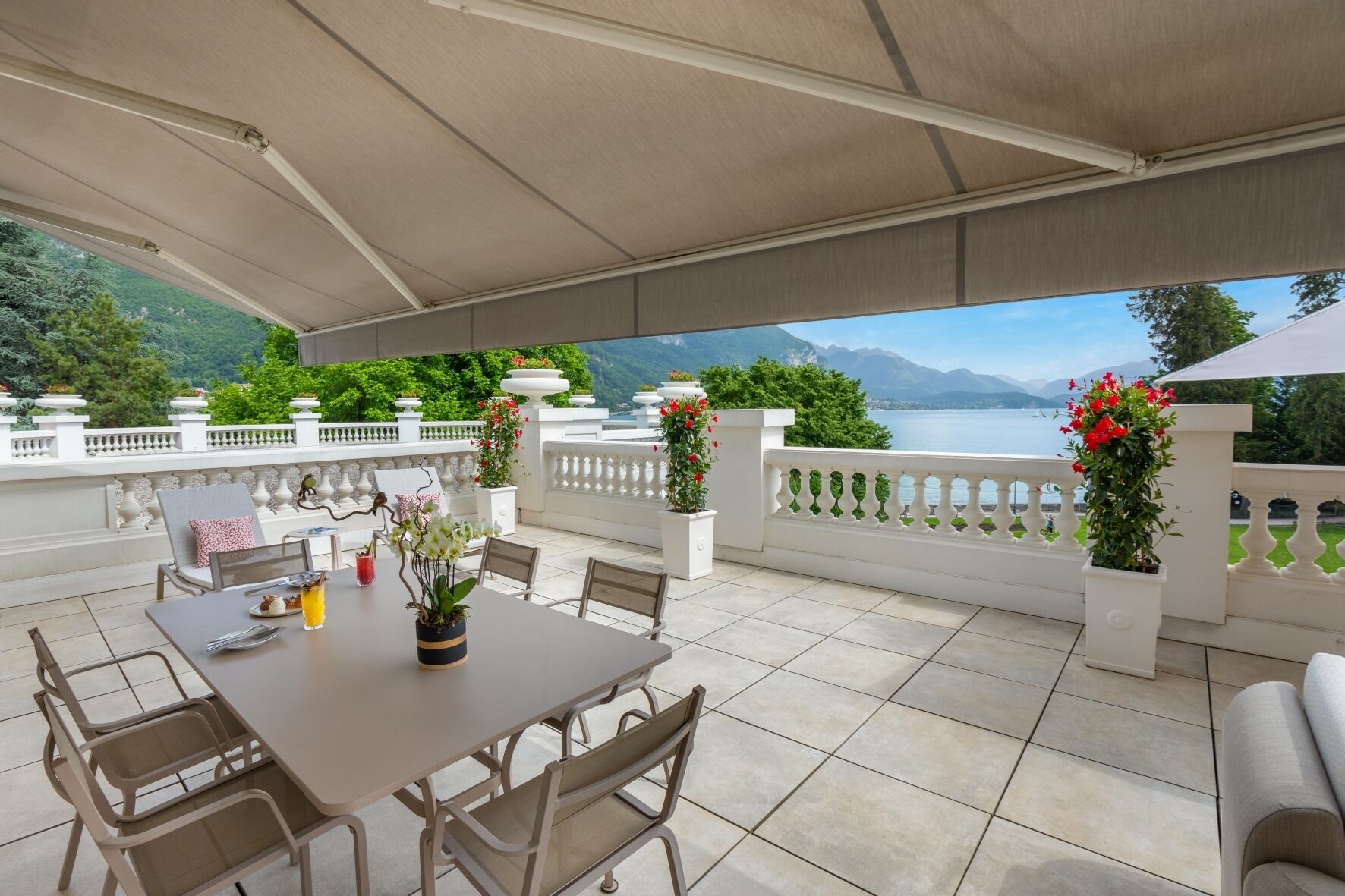 A covered outdoor patio with a table and chairs, potted plants, and a view of a lake and mountains.