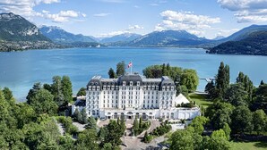 Un hotel majestuoso con una fachada simétrica, amplias ventanas y una bandera en la parte superior, rodeado de exuberante vegetación y un lago con montañas al fondo.