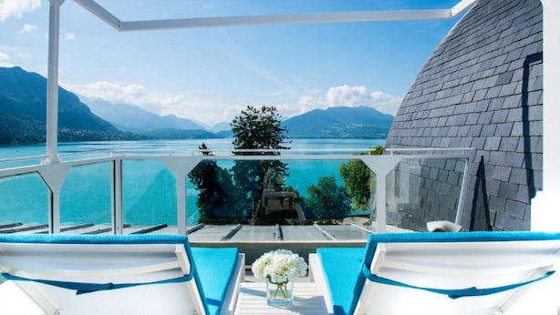 Un balcón con vistas a un lago y montañas, una mesa con flores y un edificio blanco con tejado gris.