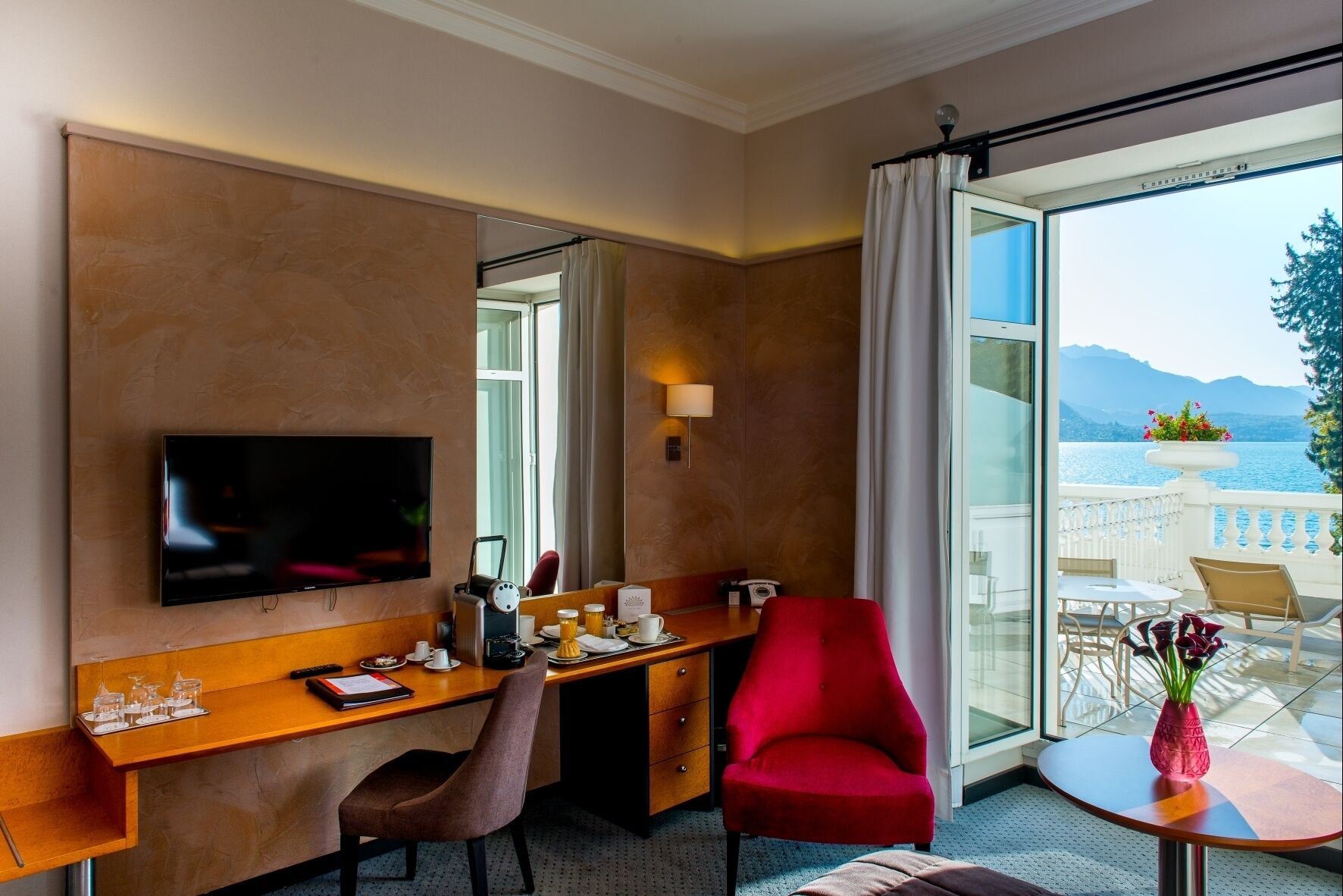 A hotel room with a TV, desk, chair, and a balcony with a view of the lake.