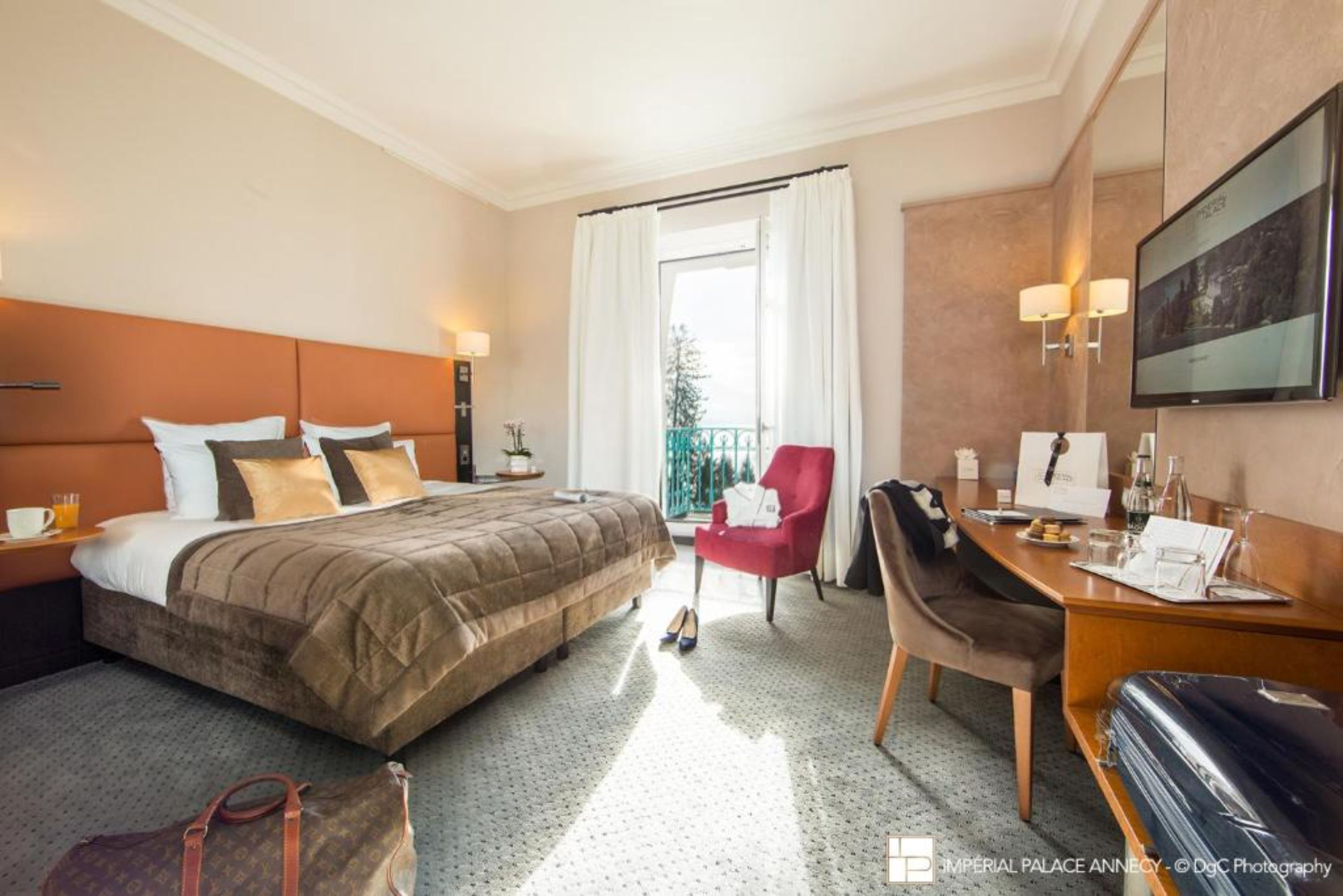 A hotel room with a bed, a desk, a chair, a suitcase, and a balcony with a view.