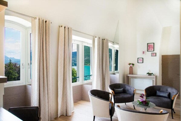 A room with a view of a lake and mountains, featuring two black leather chairs, a small table with a vase, and a fireplace with a vase of flowers. - L'Impérial Palace (Annecy)