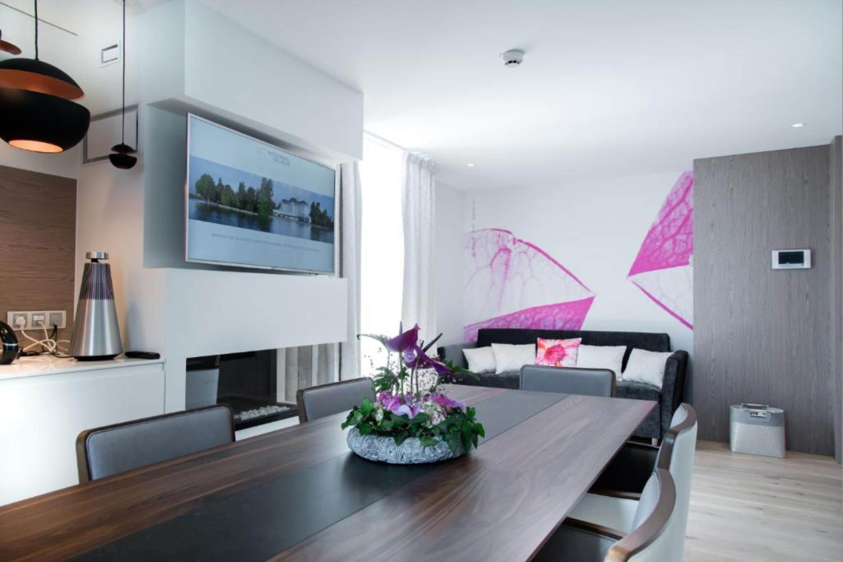 A modern dining area with a long wooden table, chairs, a flower centerpiece, a flat-screen TV on the wall, and a large window with curtains.