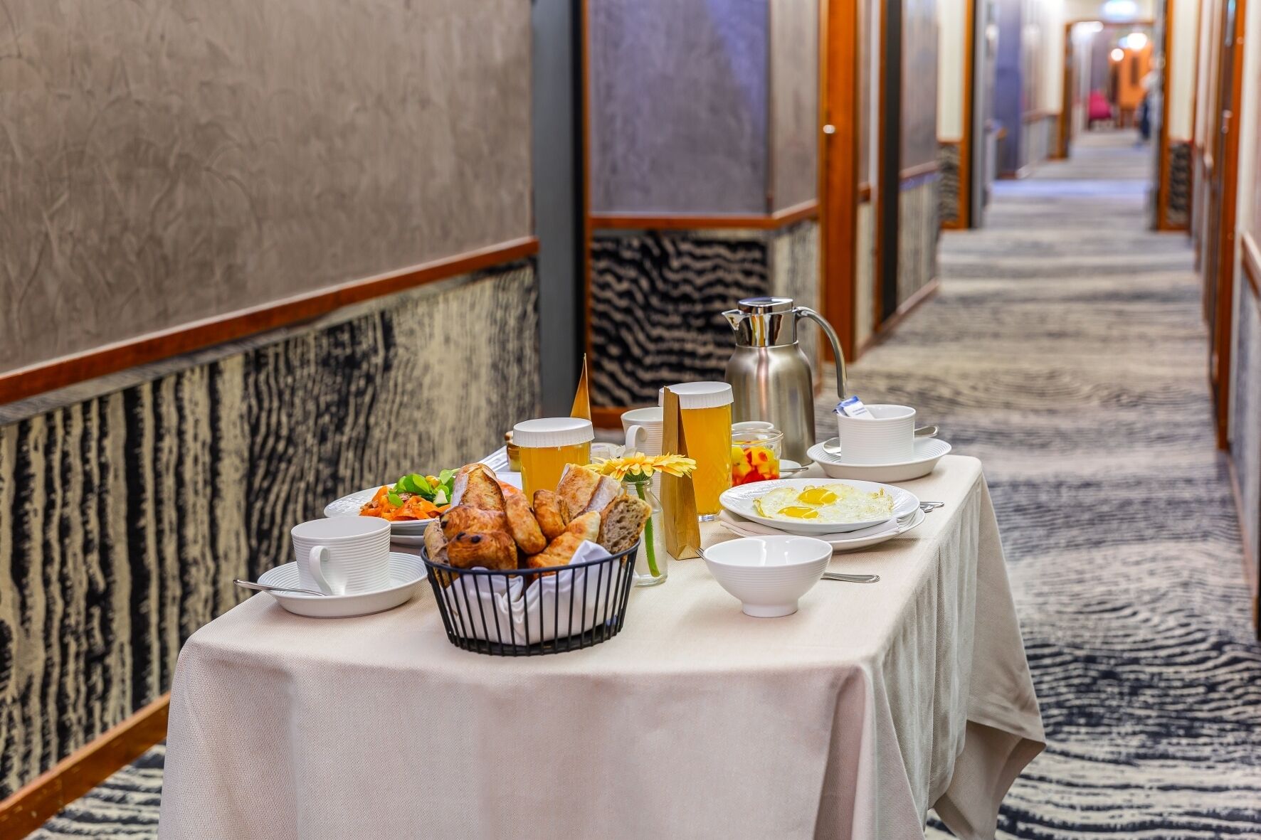 A breakfast setup with eggs, bread, and beverages on a table in a hotel corridor.