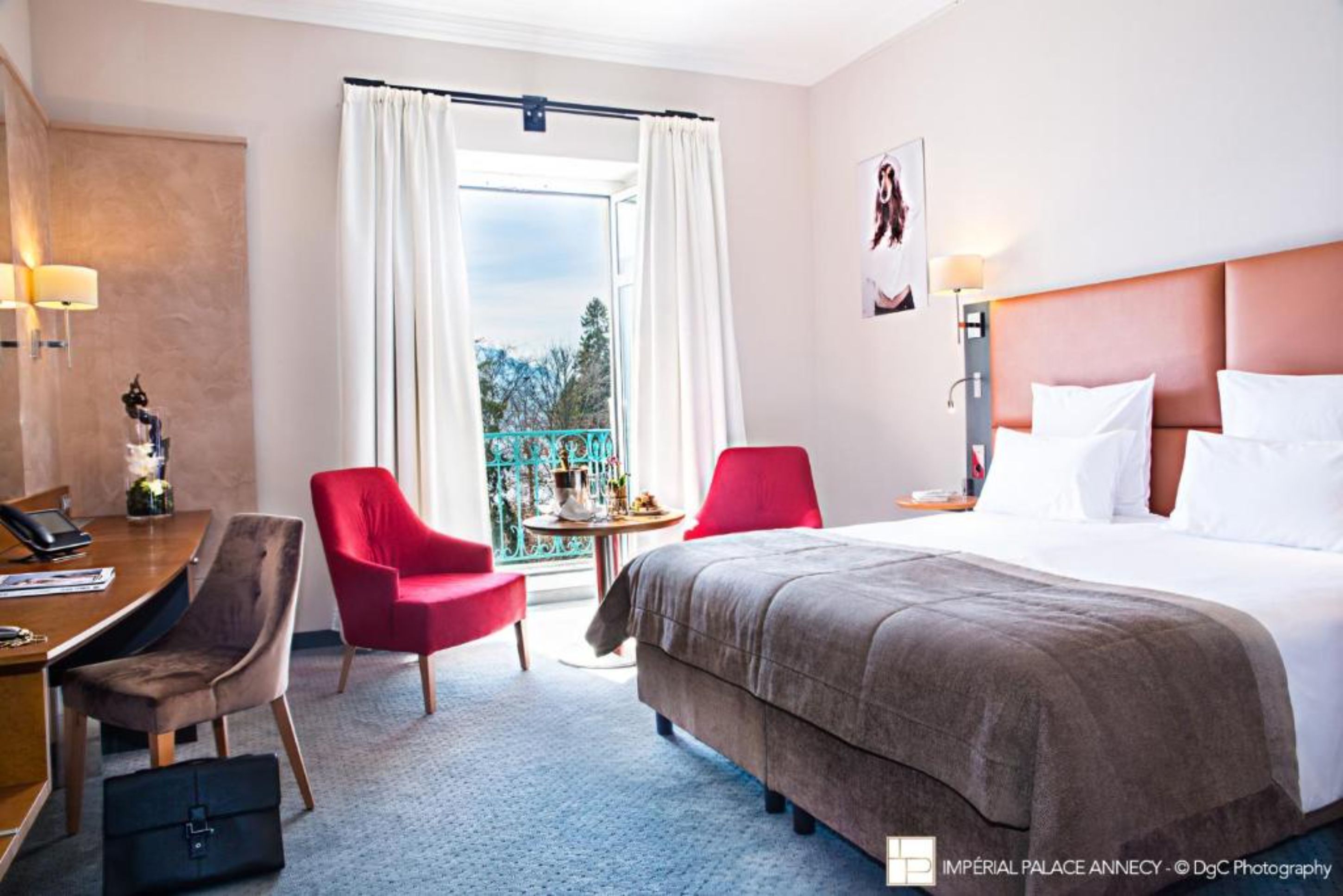 Een hotelkamer met een bed, twee stoelen, een bureau met telefoon, een tafeltje met lamp en uitzicht op het omliggende landschap.