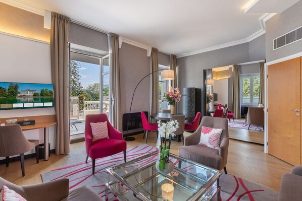 A modern hotel room with a large window, a flat-screen TV, a glass coffee table, and comfortable seating.