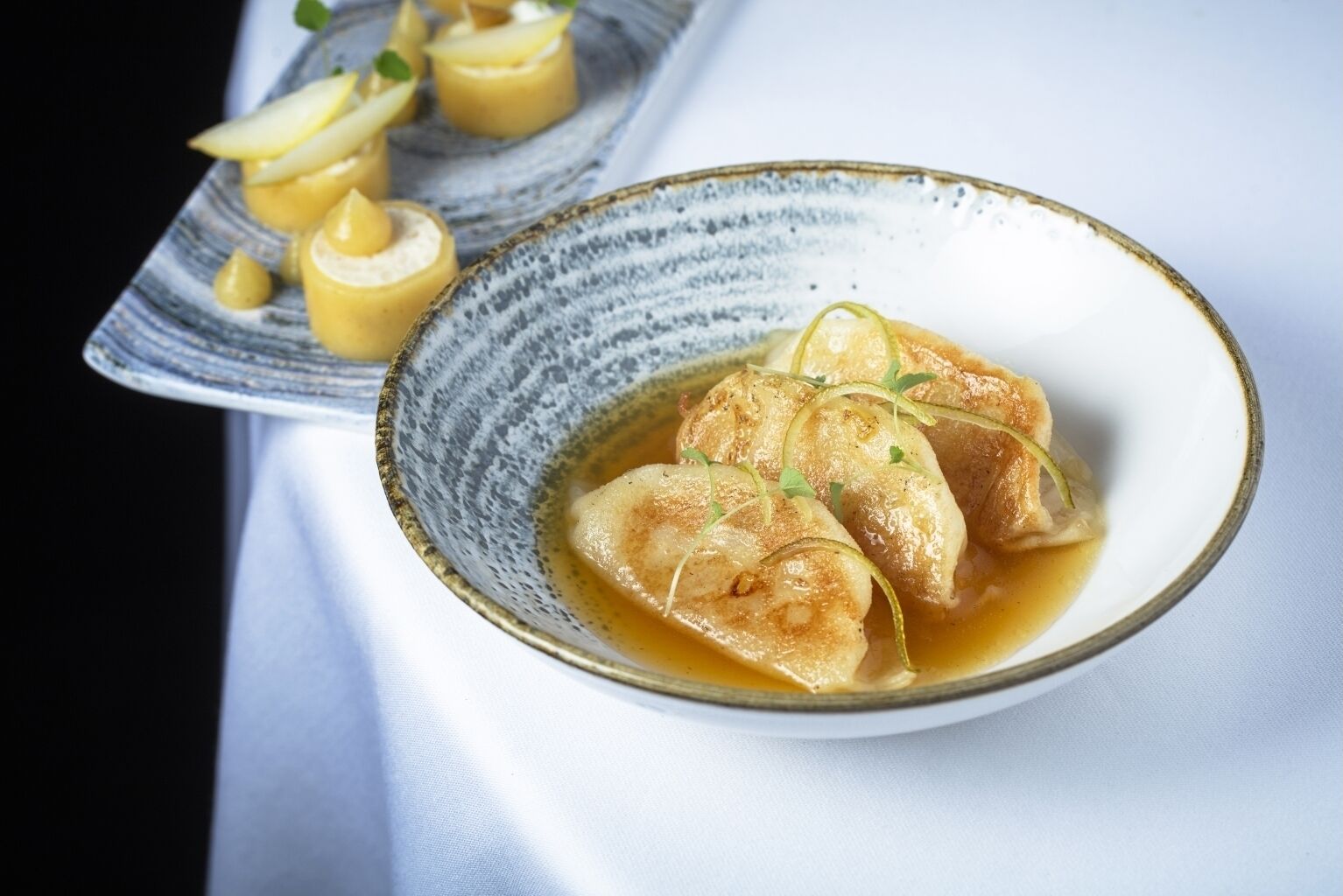 A bowl of dumplings in a ceramic dish with a sauce, garnished with herbs, and a plate with additional dumplings and a sauce container in the background.