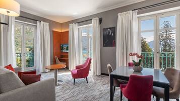 A hotel room with a sofa, chairs, a table, and a TV. There are large windows with curtains and a balcony with a view of trees and mountains.