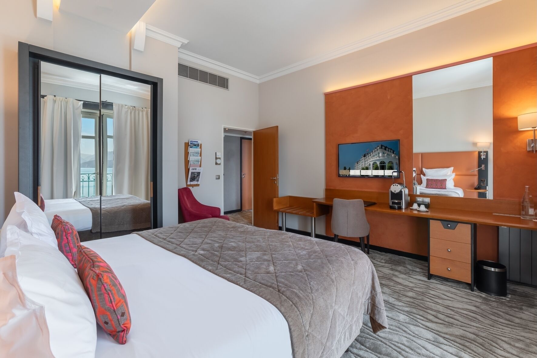 A hotel room with a large bed, a desk, a chair, a mirror, and a television.