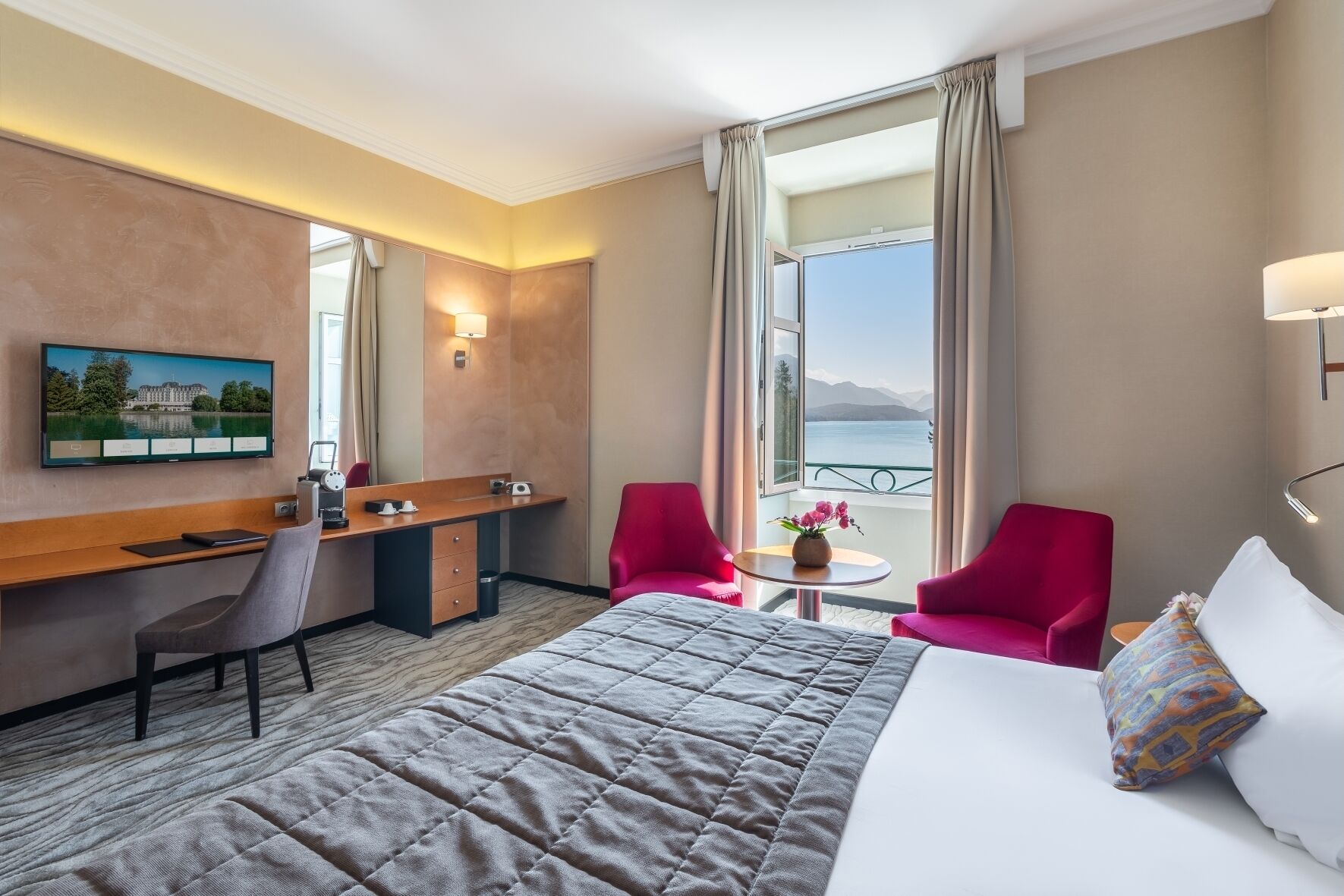 A hotel room with a bed, a desk, two red chairs, a small table with a vase, and a TV on the wall.