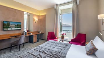 A hotel room with a bed, a desk, two red chairs, a small table with a vase, and a TV on the wall.