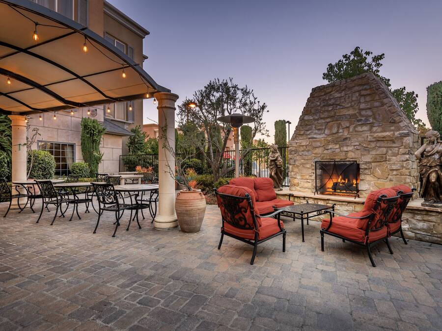 An outdoor patio with stone flooring, red upholstered chairs, a stone fireplace, and a pergola with string lights.